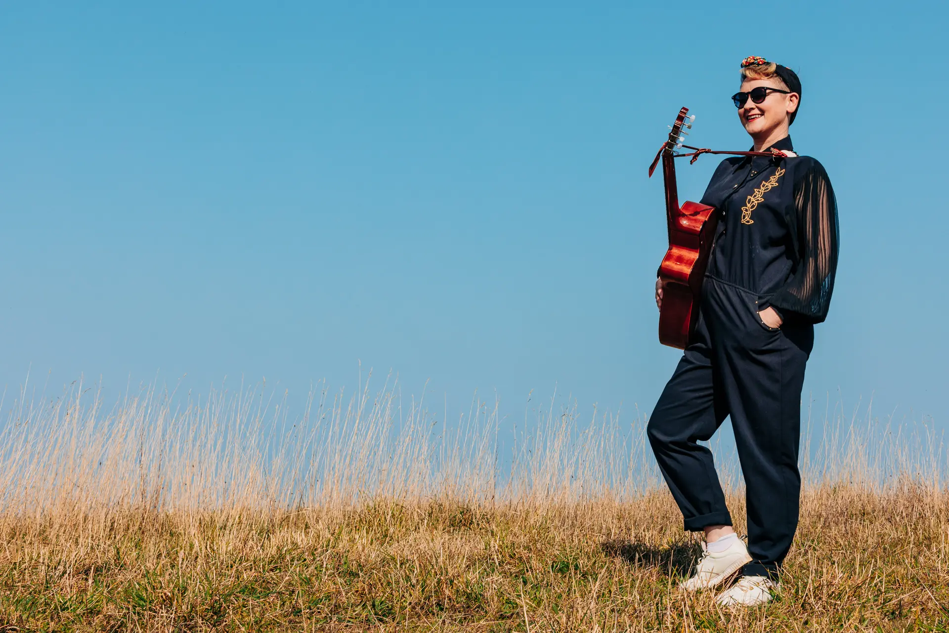 Image shows a woman dressed in black standing with a guitar on the edge of a cliff with bright blue sky behind her. Shot for the promotion of Bertie Anderson's Poppyland songs performances.