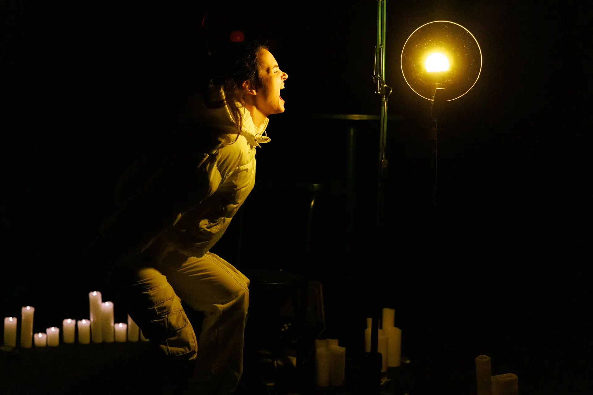 Performer dramatically screaming from the side, illuminated by a bright stage light, during 'Ghost Stories by Candlelight' by HighTide Theatre at Eastern Angles. Captured by performing arts photography specialist, Fourth Wall Photography.