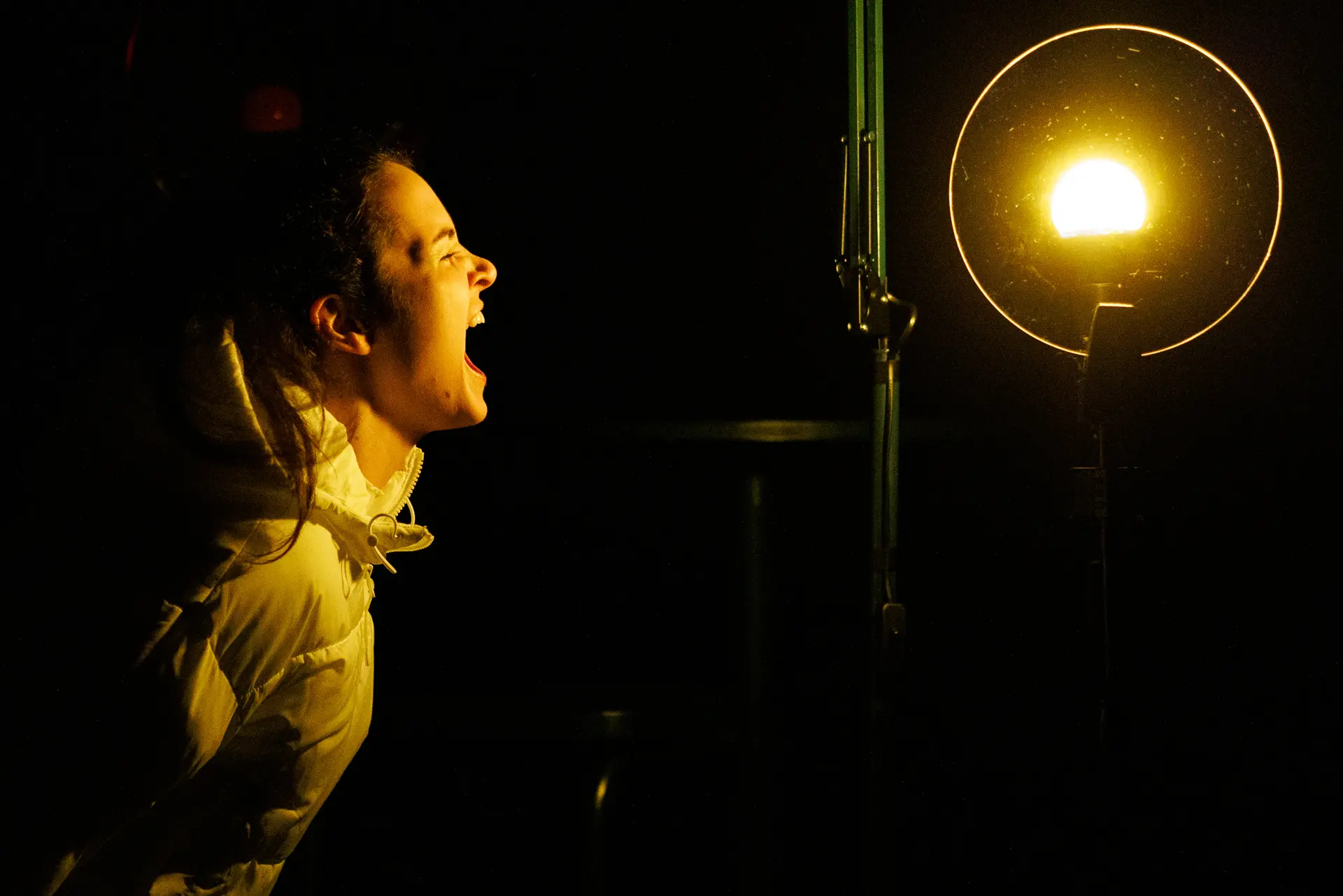 Performer dramatically screaming from the side, illuminated by a bright stage light, during 'Ghost Stories by Candlelight' by HighTide Theatre at Eastern Angles. Captured by Theatre photography specialist, Fourth Wall Photography.