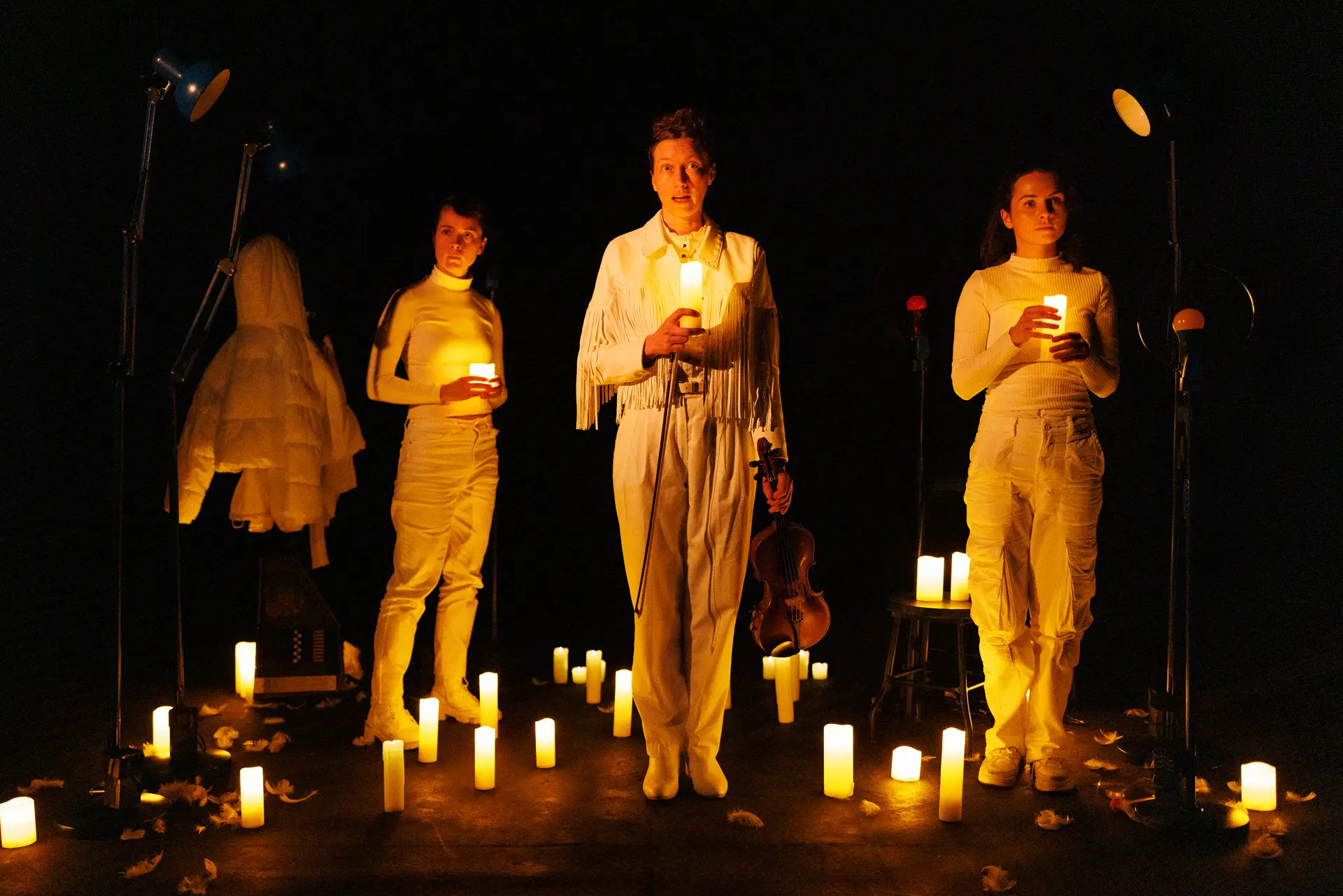 Three individuals dressed in white stand on a dark stage illuminated by numerous candles. The person in the centre holds a candle and a violin. To their left, another person in white holds a candle, next to a white hooded jacket on a stand. To the right, a third person in white also holds a candle. Captured by specialist performing arts photographer Fourth Wall Photography