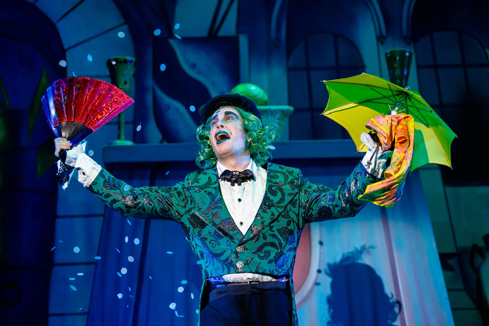 Fleshcreep from the Jack and the Beanstalk pantomime at the Marina Theatre in Lowestoft, with green hair, holding a red fan and a green umbrella with falling confetti. Captured by Theatre photography specialist, Fourth Wall Photography.