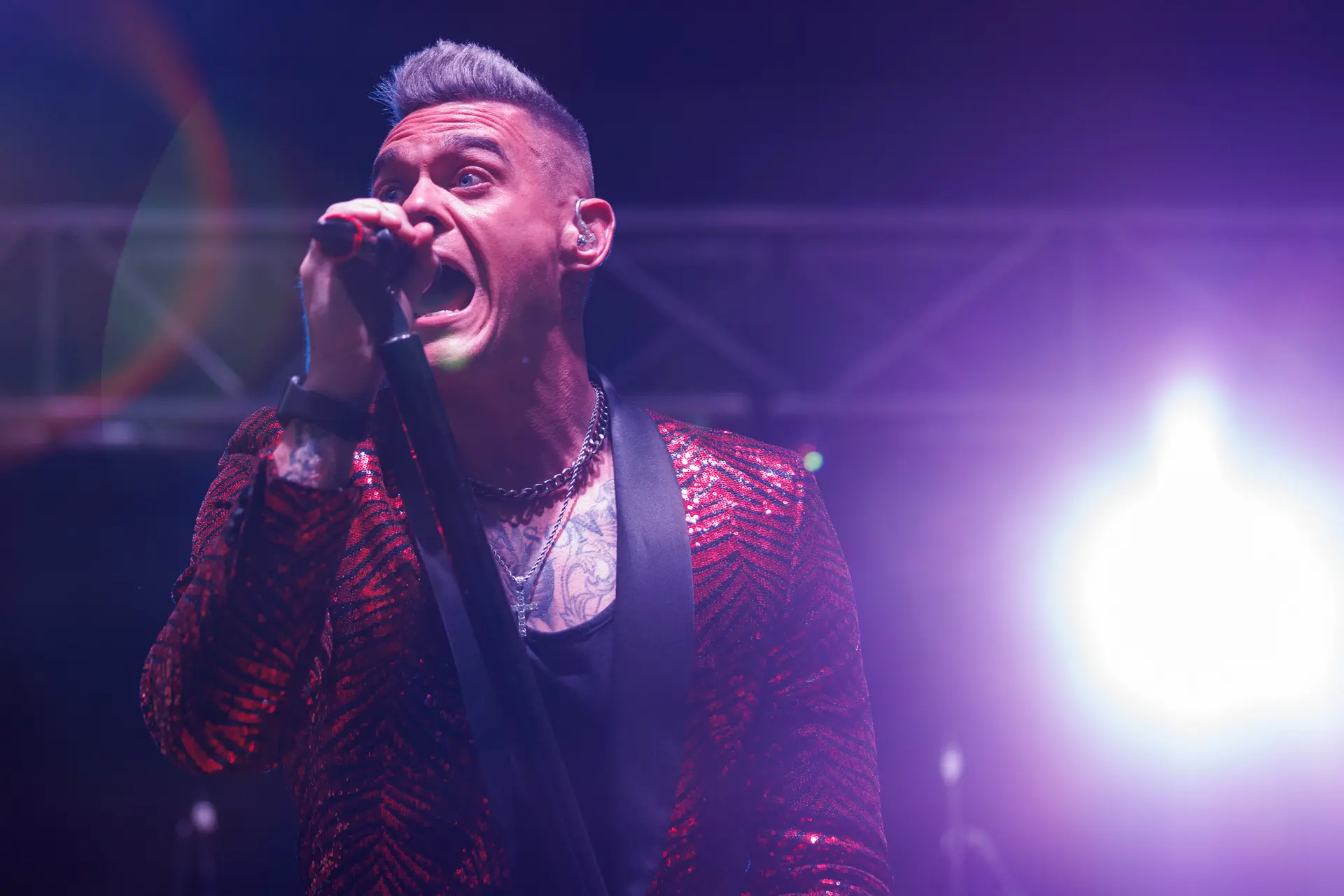 Dan Budd performing as Robbie Williams at the Legends Festival in Cromer, wearing a red sequin jacket and singing intensely under purple stage lighting. Captured by Live Music photography specialist, Fourth Wall Photography.