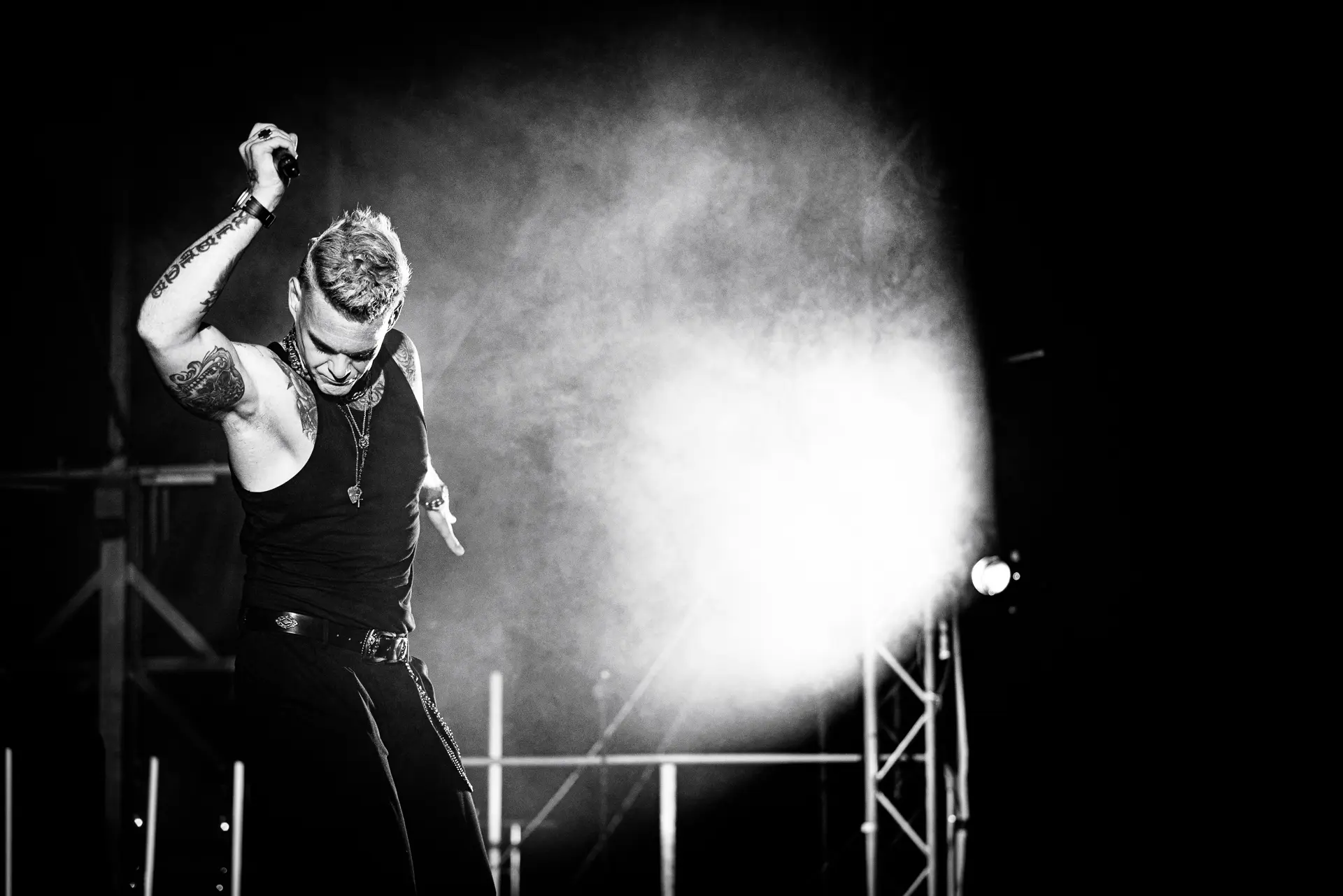 Black and white image of Dan Budd performing as Robbie Williams at the Legends Music Festival in Cromer, raising his arm and singing under a dramatic spotlight and haze. Captured by Live Music photography specialist, Fourth Wall Photography.