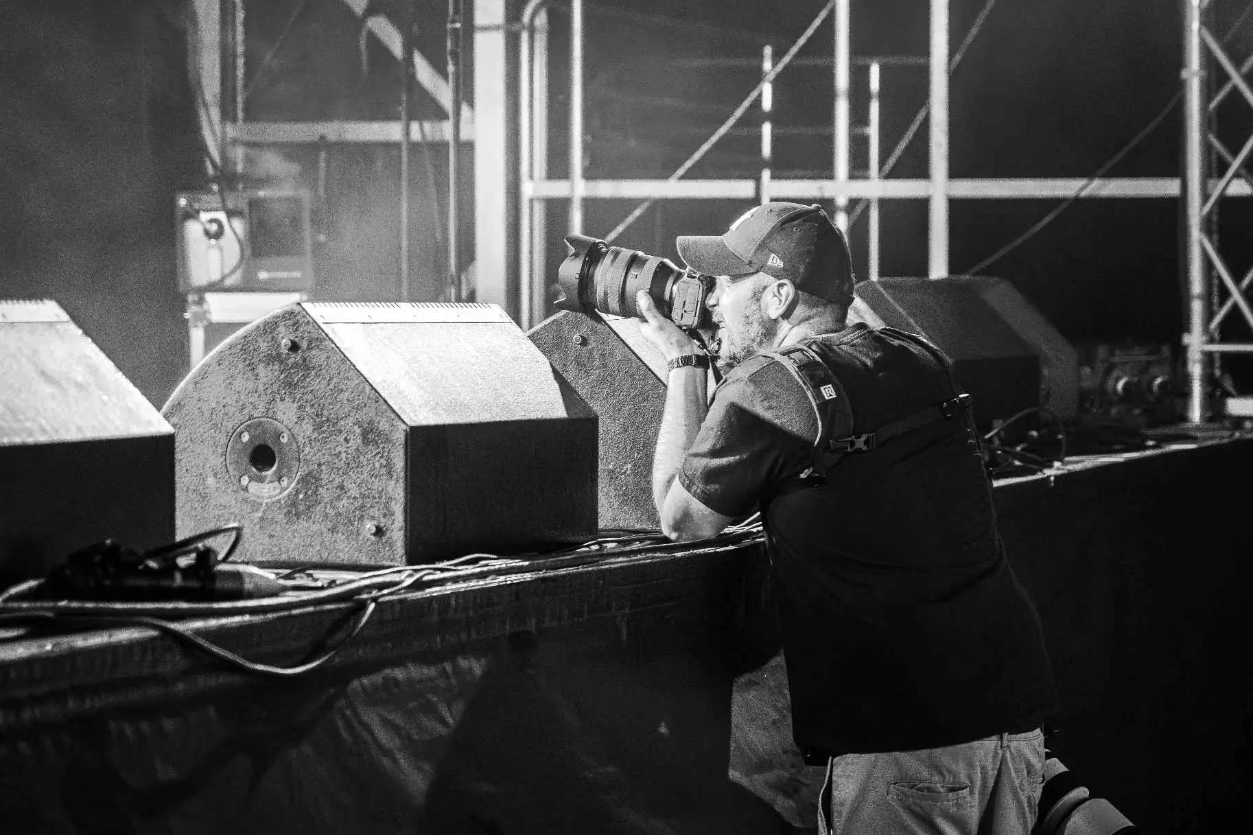 Image of Matt Coomber-Such a performing arts photographer in a cap and harness leans against stage speakers, aiming a professional camera during a live performance.