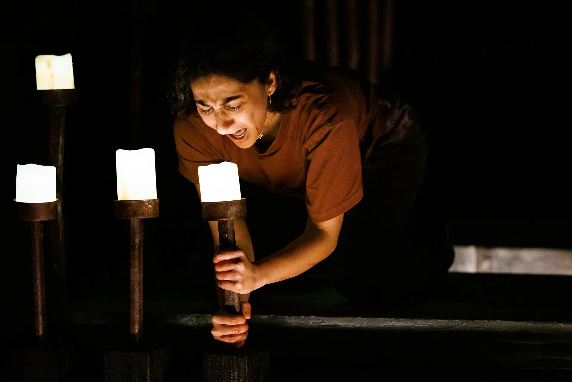 A theatrical shot of a performer screaming on a dark stage. They are wearing a rust-coloured top and dark pants and are screaming into a candle. The stage is dimly lit by square, glowing candles on tall, slender posts and shorter stands surrounding the performer. Image is from More... Ghost Stories by Candlelight by HighTide theatre performed at the Eastern Angles Centre, Ipswich.