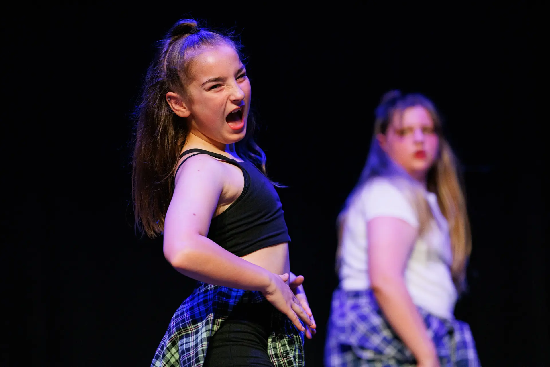 Young female dancer performing with an expressive face and energy on a dark stage, another dancer blurred in background. Dancers performing as part of the North Suffolk Schools Dance Festival the the Marina Theatre in Lowestoft. Captured by Dance photography specialist, Fourth Wall Photography.