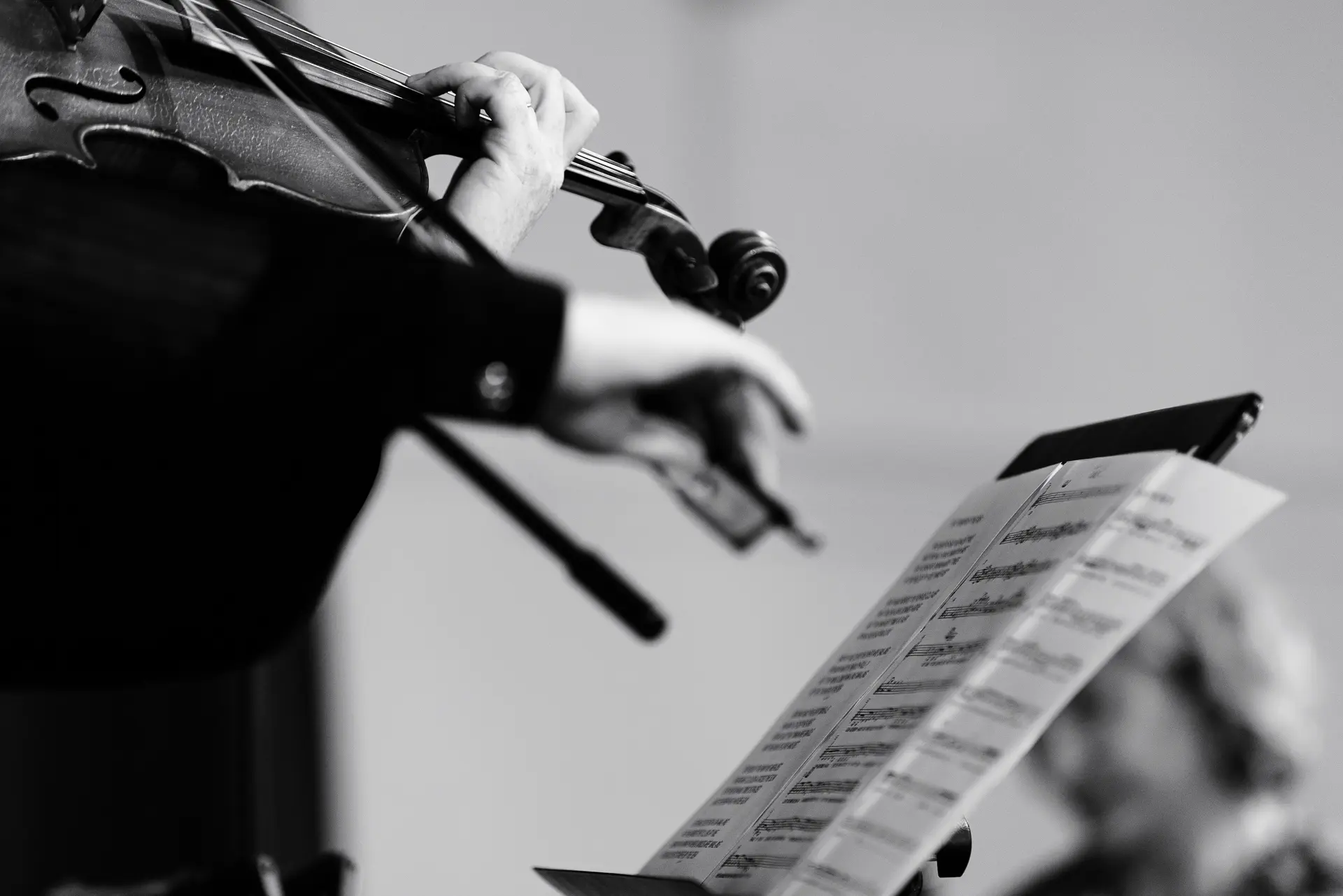 A close-up, black and white image focusing on the hands of a musician playing a violin, with sheet music on a stand visible in the foreground. Captured by live music photography specialist Fourth Wall Photography